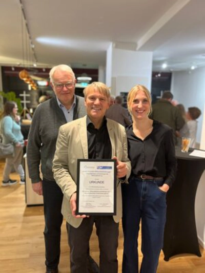 drei Personen lächeln in die Kamera. Eine hält eine Urkunde in der Hand.